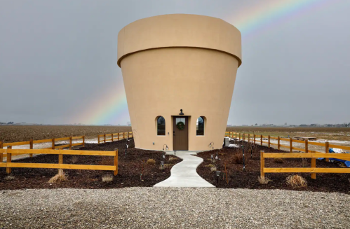 A unique round building resembling a large pot, set in a field with a rainbow in the background and a path leading to it.